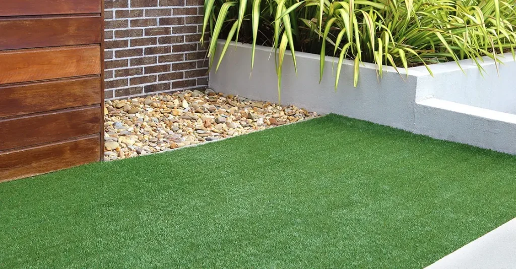 Green artificial grass area next to stone pebbles and tall plants against a wall