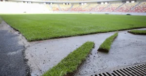 Grass being laid down on a sports field, with uneven patches and a drainage grate visible
