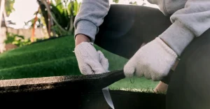 A person in gloves cuts material with a knife while kneeling on grass