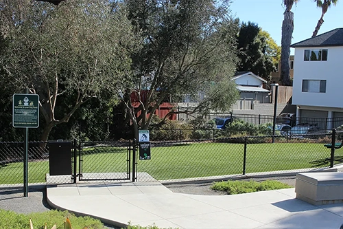 Park entrance with a fenced grassy area, trees, and nearby buildings