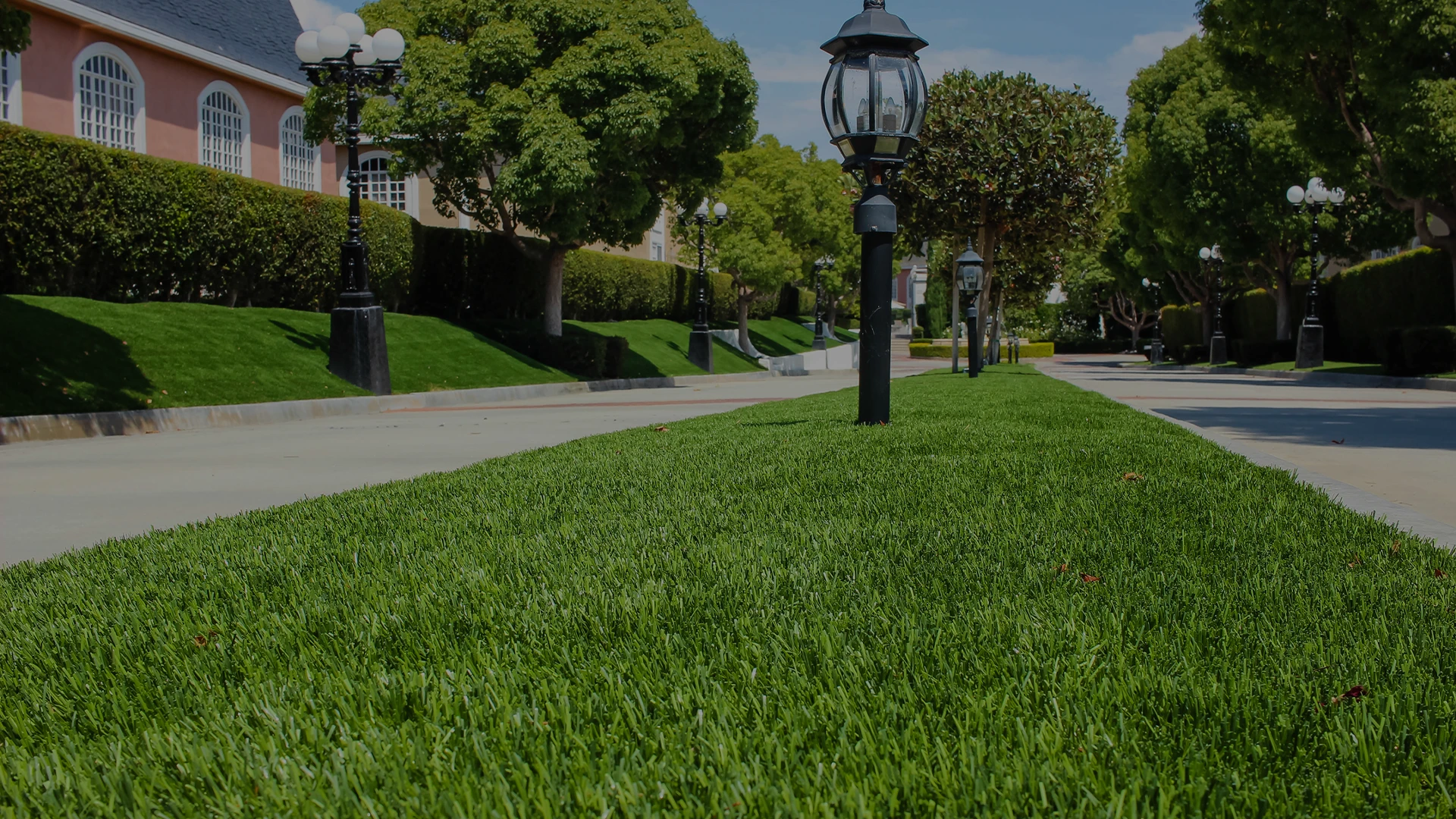 Lush green grass lined with street lamps beside a paved walkway and trees