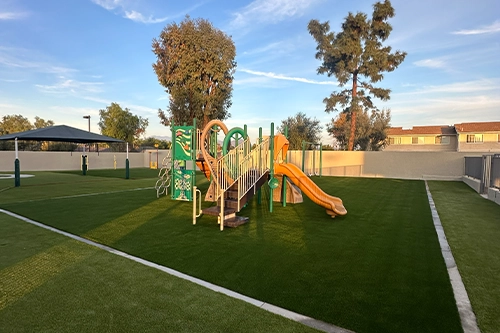 Playground with a slide and climbing structure on grass, surrounded by trees and buildings