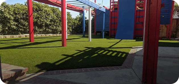 Playground area with green grass, red and blue structures, and trees in the background