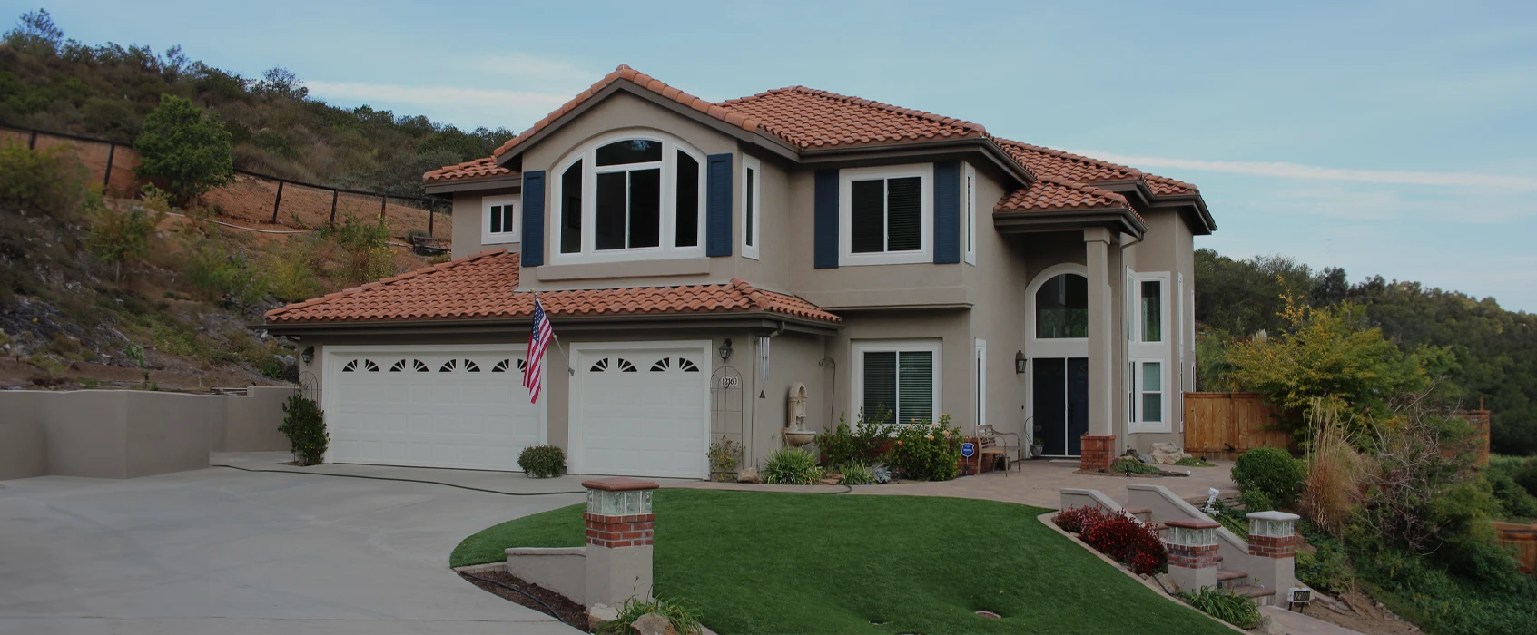 Two-story house with a tiled roof, green lawn, and driveway, surrounded by hills