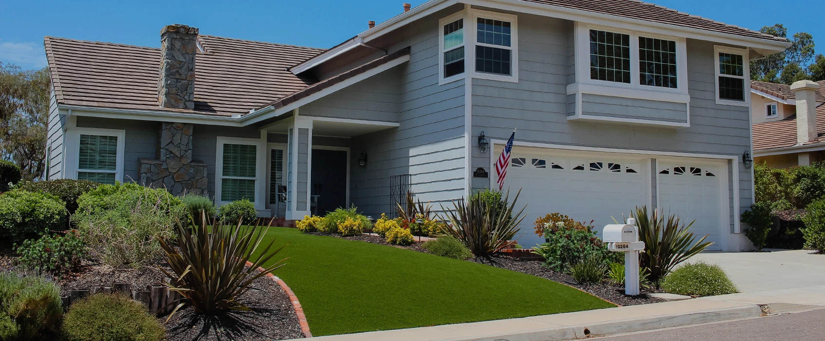 Light gray house with a front lawn and flowerbeds, two-car garage, and blue sky