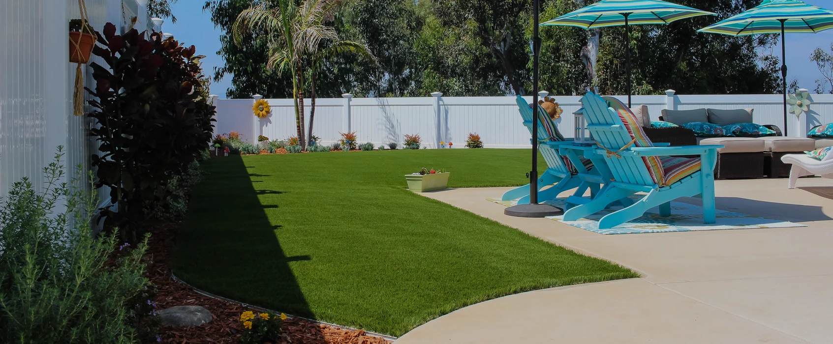 Green lawn with a patio, blue chairs, umbrellas, and plants in a backyard setting