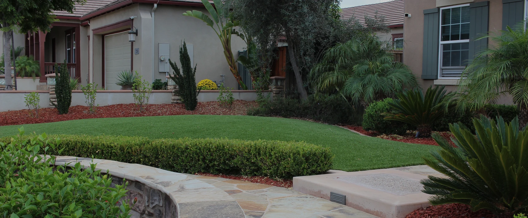 Front yard view with green lawn, shrubs, and stone pathway near residential buildings
