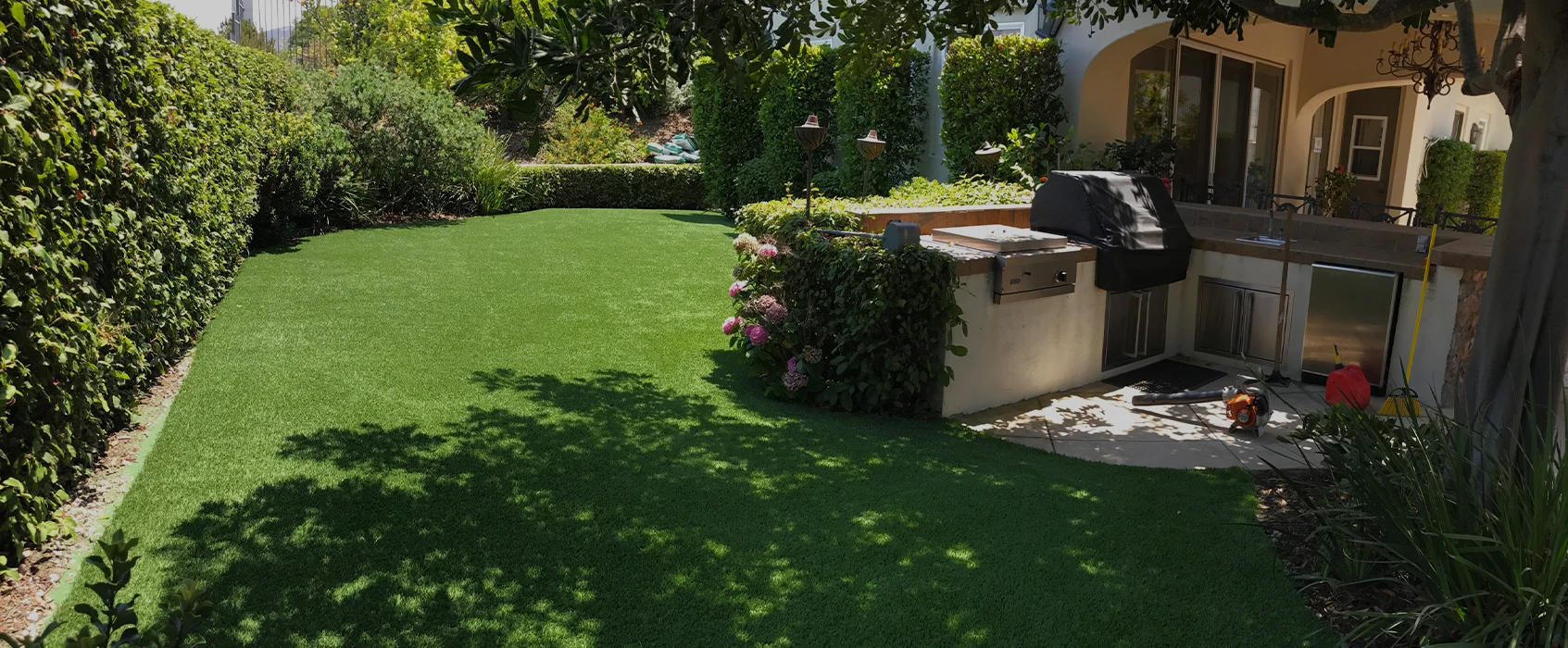 Backyard view with green artificial grass, shrubs, and a grill near a patio