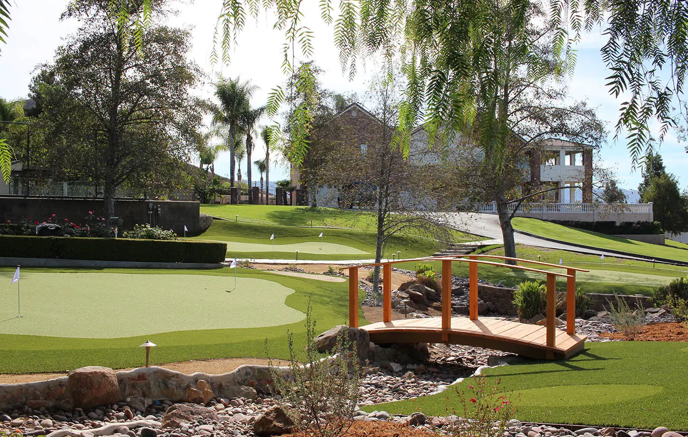A landscaped area with a golf green, a wooden bridge, trees, and a building in the background