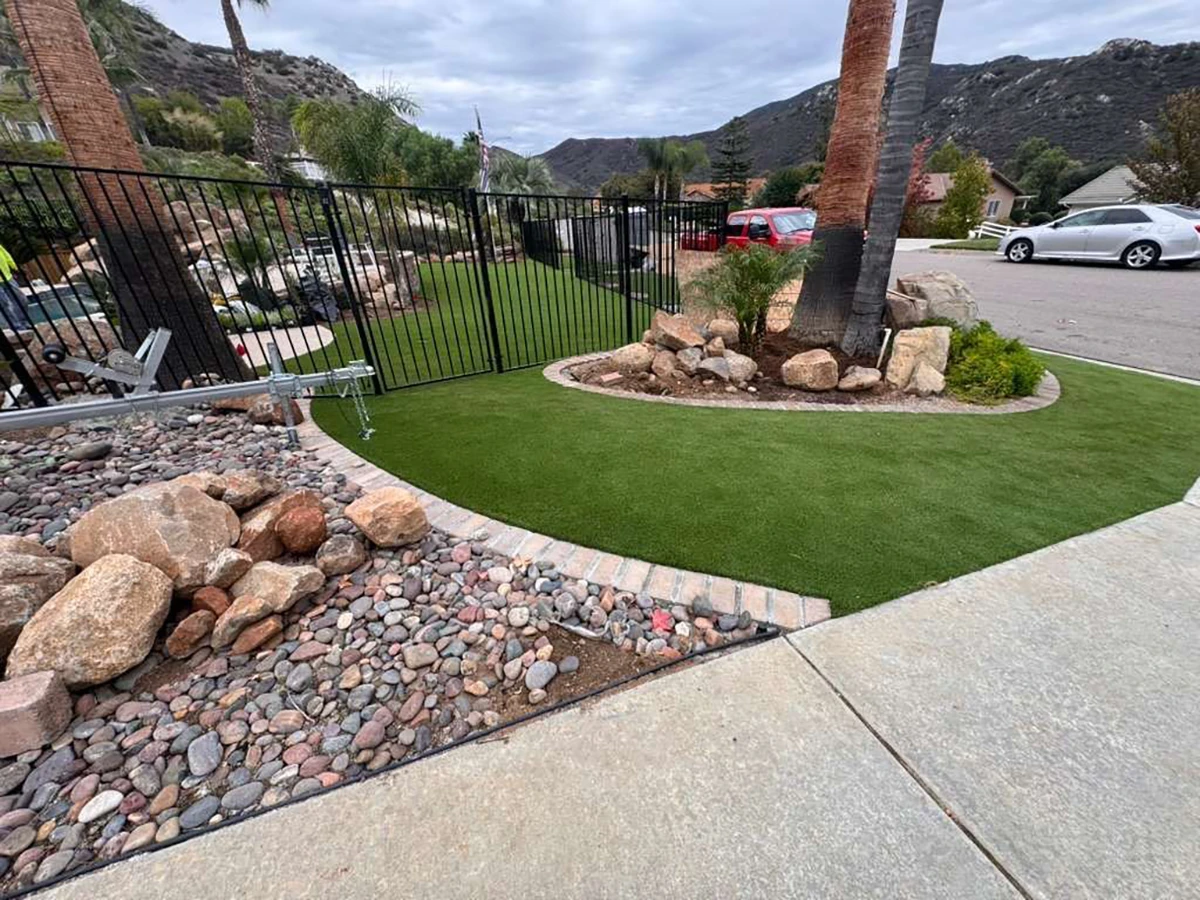 A landscaped area with a fence, grass, rocks, and palm trees near a driveway