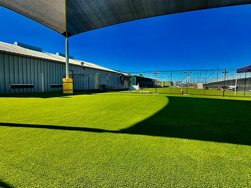 Green artificial turf under a shaded area, with buildings and a fence in the background
