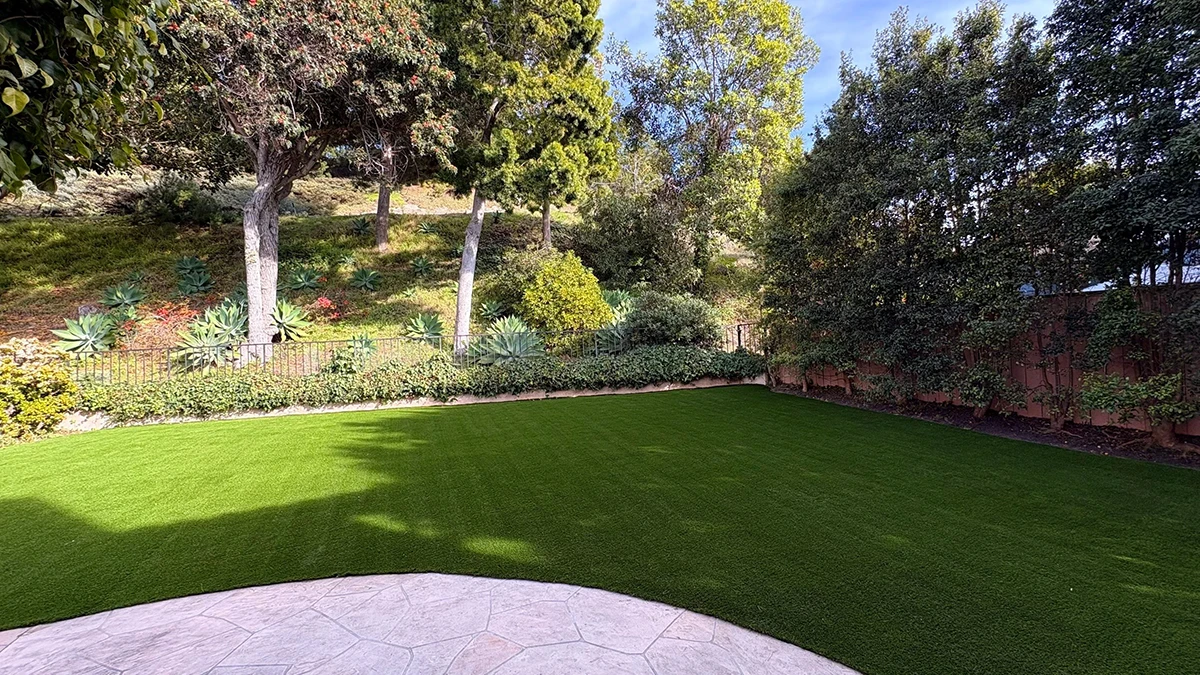 View of a grassy lawn bordered by trees and bushes, with a stone patio in the foreground