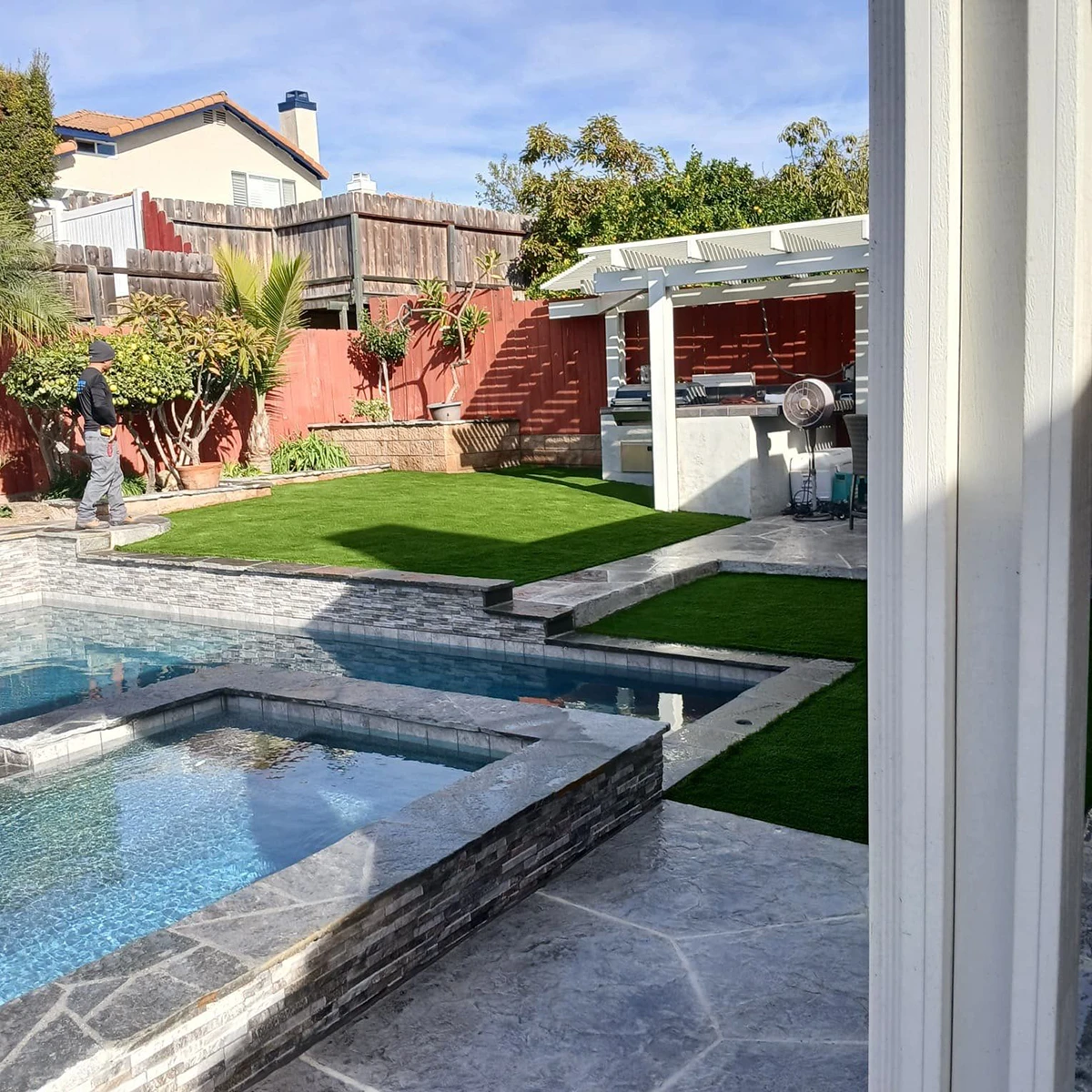 Backyard with a pool, artificial grass, and a patio area. A person is walking on the stone path