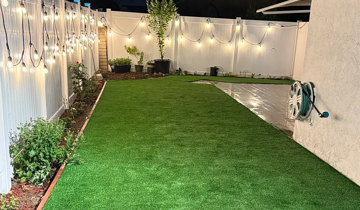 A backyard with artificial grass, plants, and string lights on a white fence