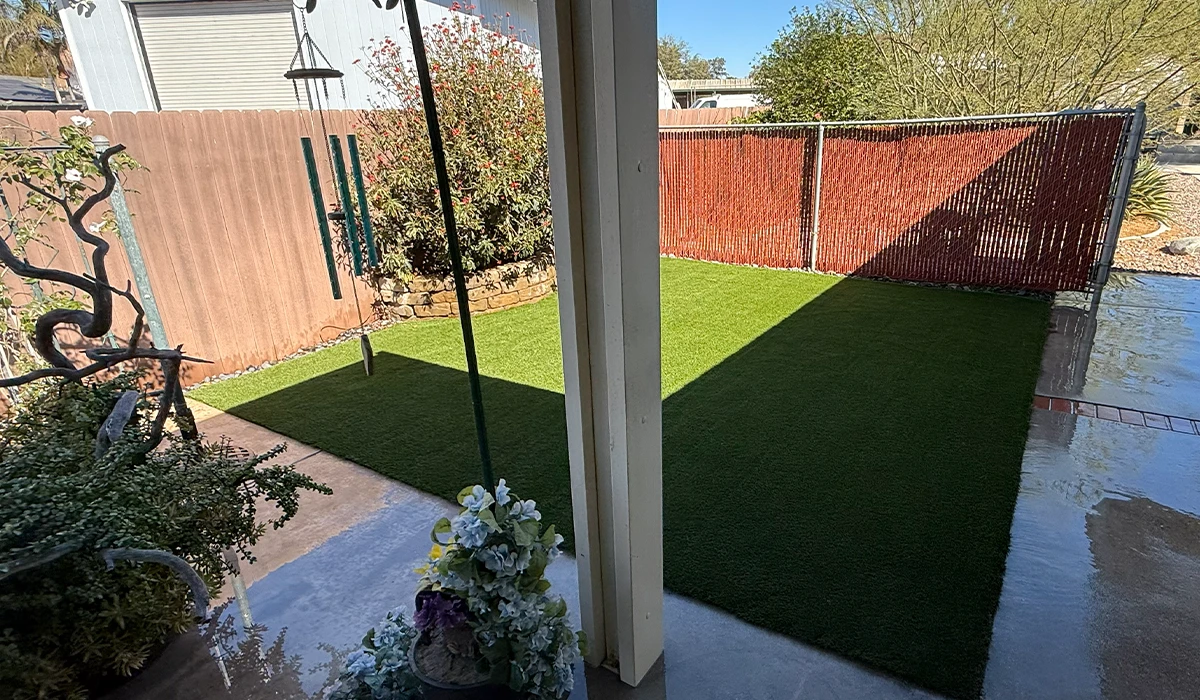 View of a grassy area enclosed by a fence, with a patio and some vegetation in the foreground