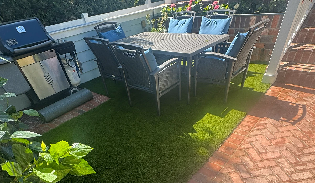Patio with a table and chairs, a grill, and artificial grass. Flowers in the background