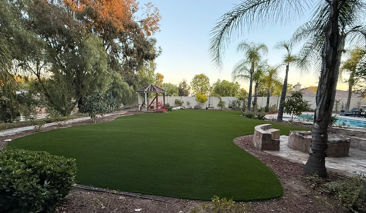 A green lawn surrounded by trees and a play structure, with a pool in the background