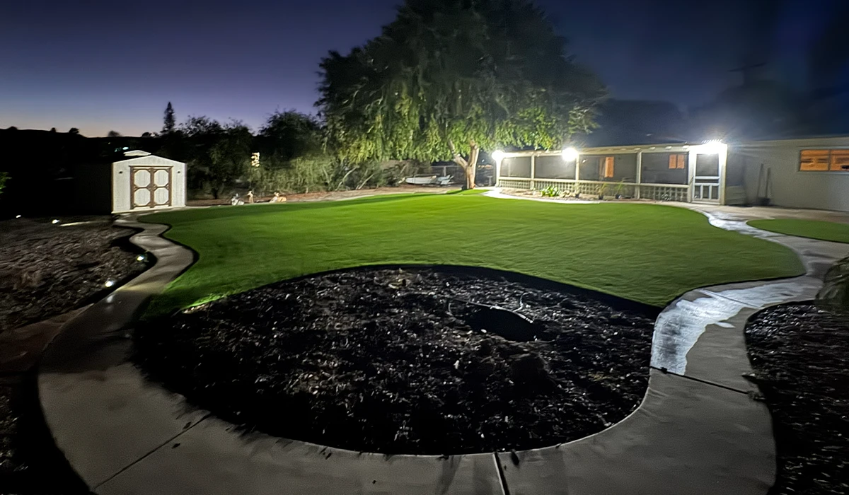 Well-lit yard at night with a grassy area, circular flower bed, and a path