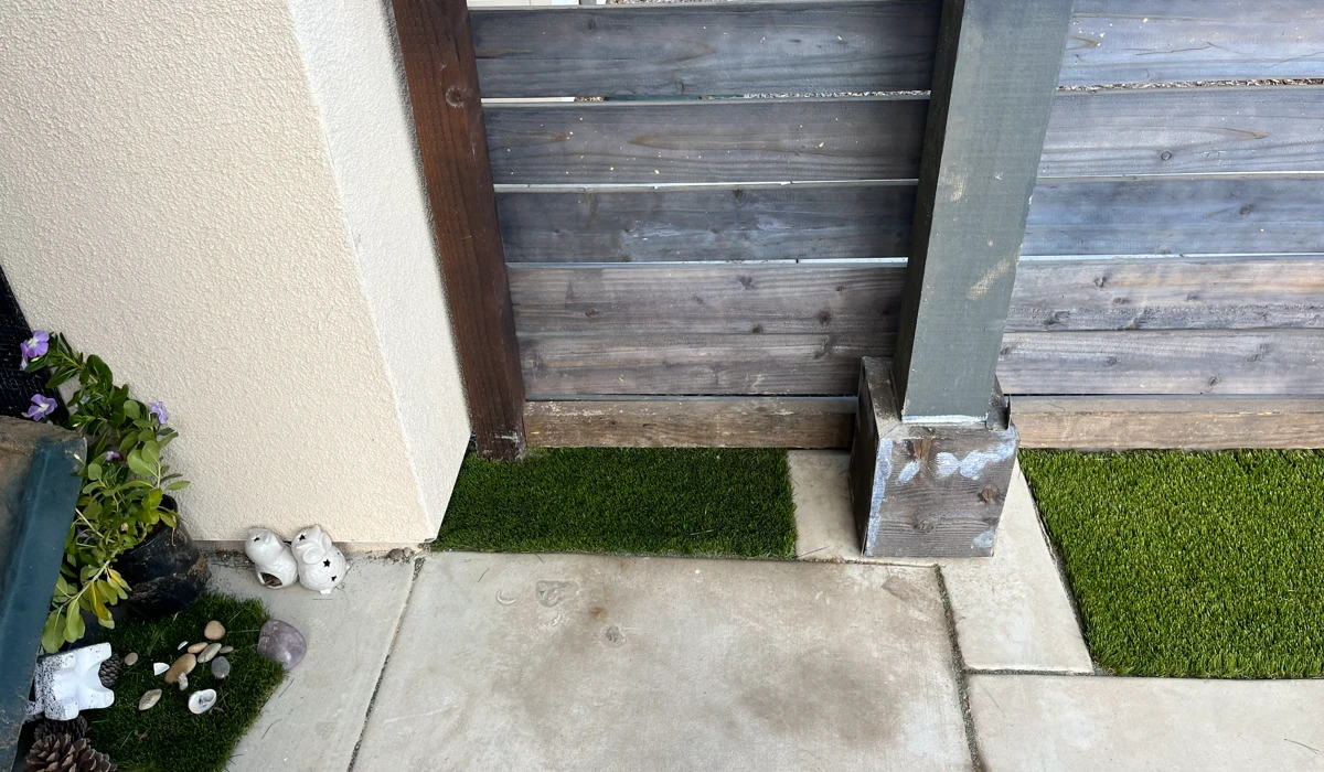 A concrete pathway with artificial grass on the sides and a wooden fence in the background