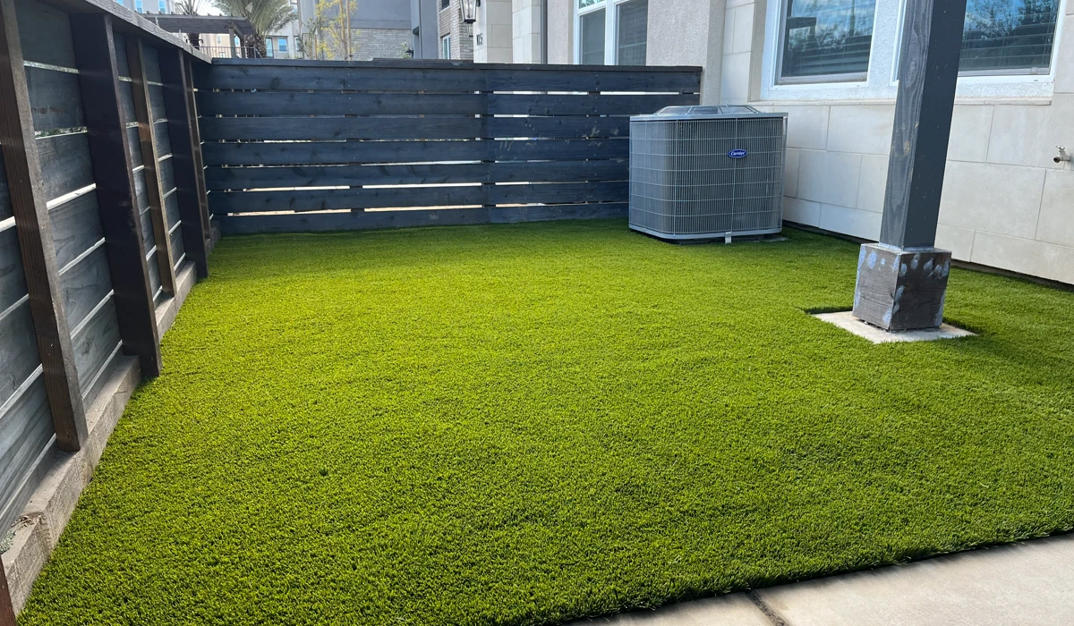 A small yard with artificial grass, wooden fence, and an air conditioning unit