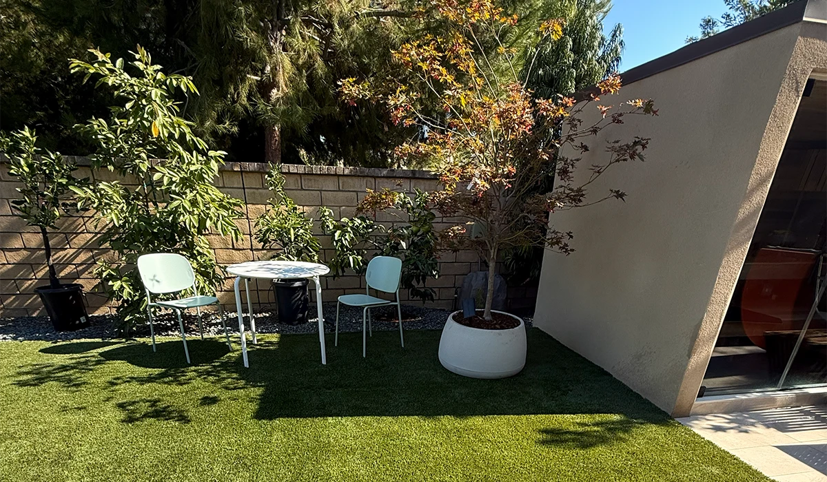 A patio with a table, two chairs, a planter, and a small tree, surrounded by grass