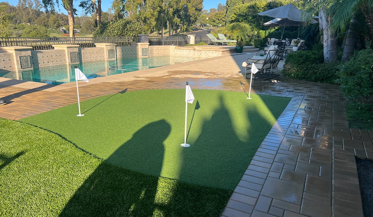 A small putting green with flags, near a pool and patio area with trees