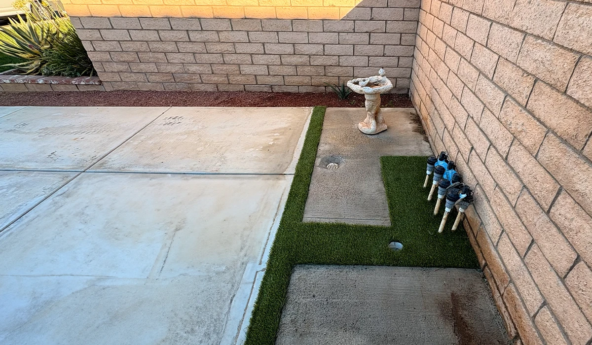 A patio area with artificial grass, a birdbath, and sprinkler heads along a brick wall