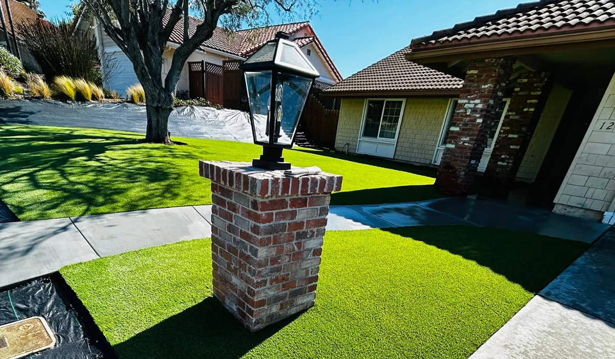A brick lamp post with a lantern stands on green grass near a house