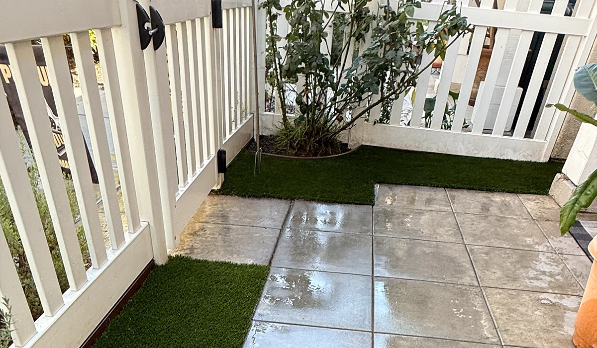Patio area with green artificial grass and tiled section beside a white fence and plants