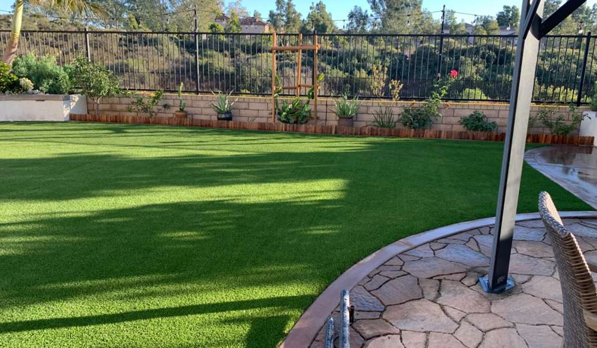A green artificial turf lawn next to a stone patio, surrounded by plants and a fence