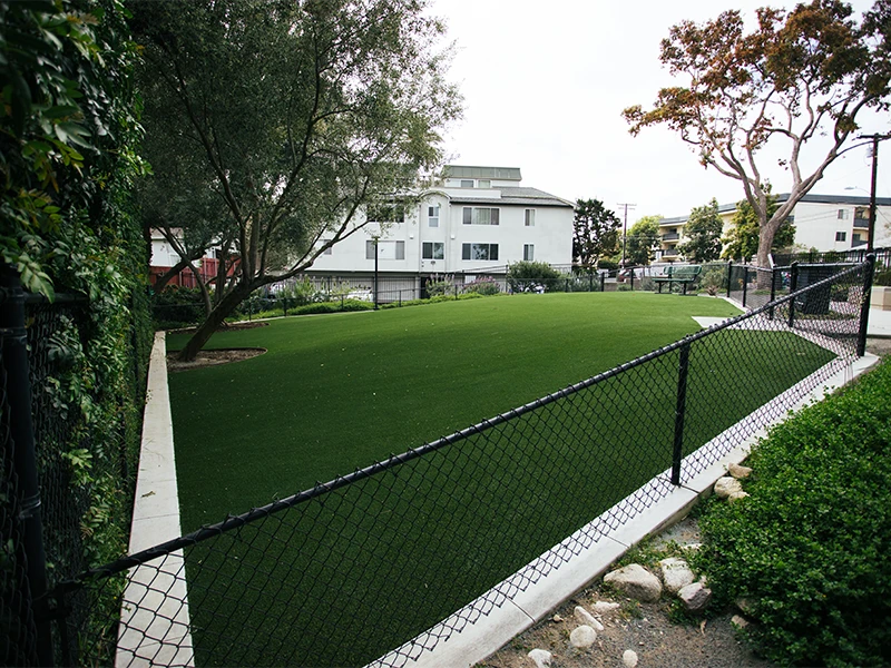 A green artificial turf area surrounded by a fence and trees, with a building in the background