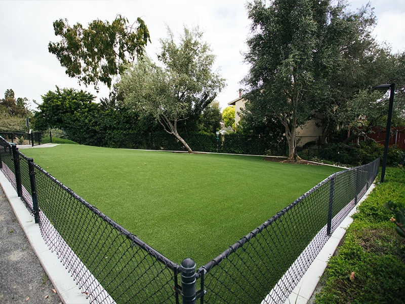 Green artificial turf enclosed by a black fence, surrounded by trees and shrubs