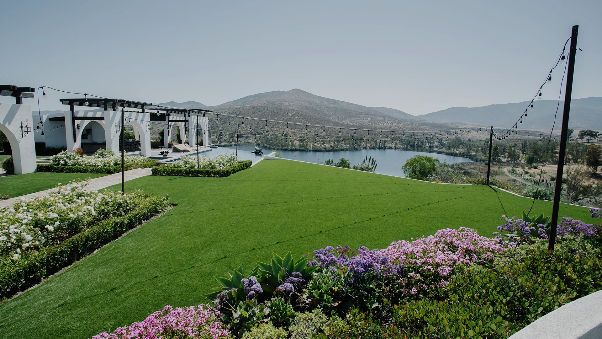 A green lawn with flower beds overlooks a lake and mountains in the distance