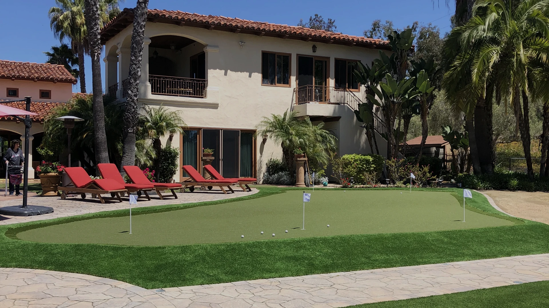 A house with a putting green, lounge chairs, and palm trees in a sunny area