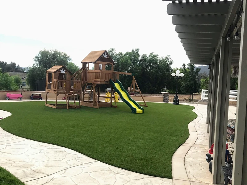 Playground with wooden structure, slide, and artificial grass. Trees in the background