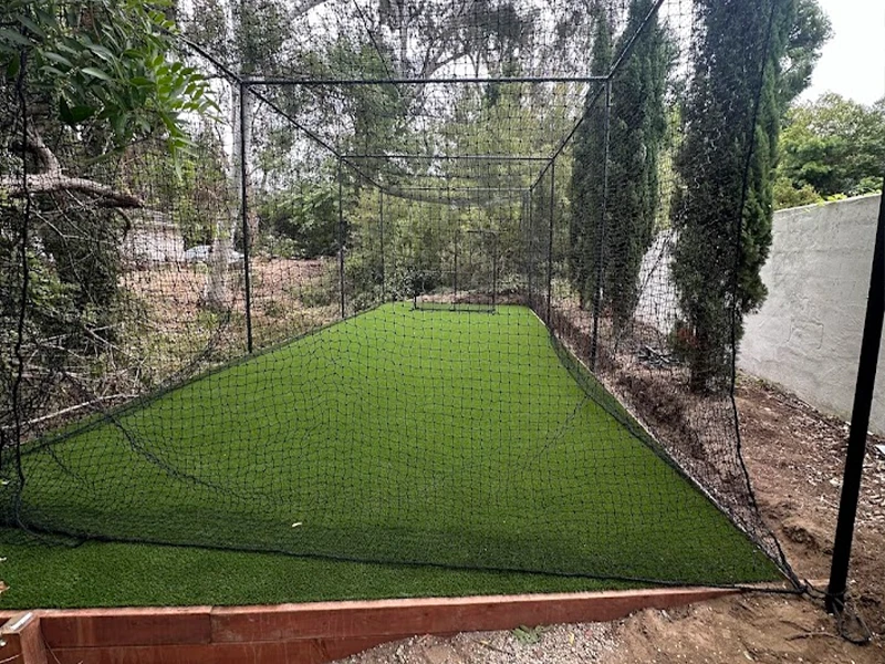 A netted batting cage with artificial grass, surrounded by trees and a wall