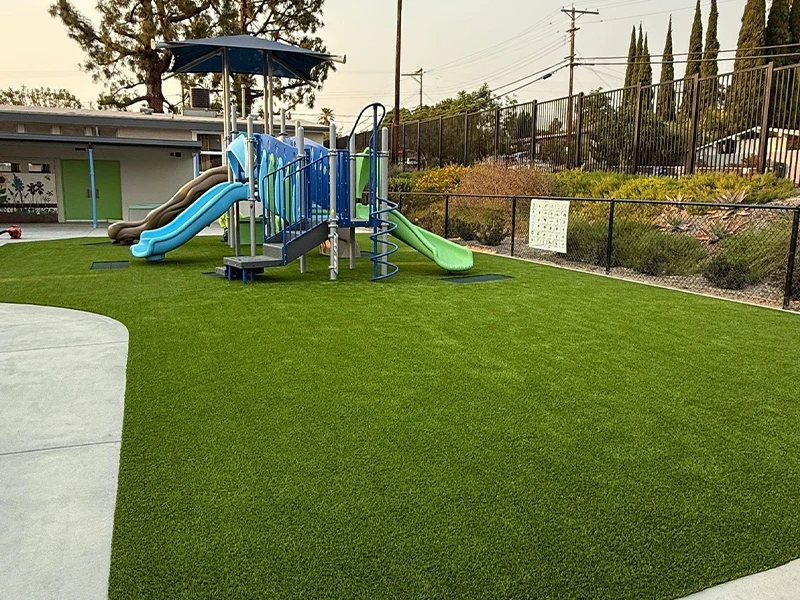 Playground with blue and green slides on artificial grass. Fenced area with buildings in background